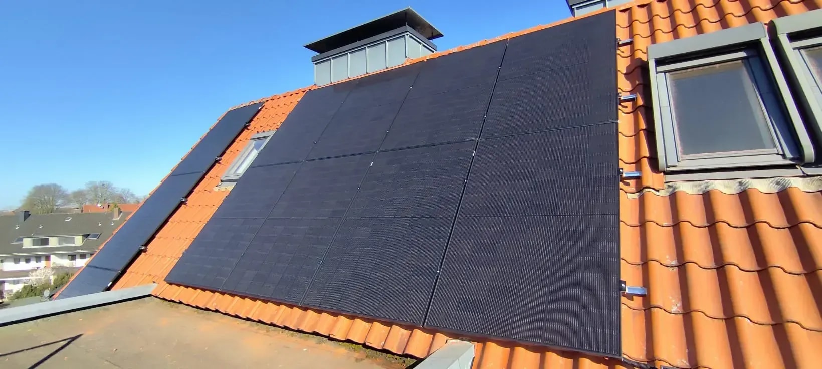 Photovoltaikanlage mit schwarzen Solarmodulen auf einem Ziegeldach unter blauem Himmel.